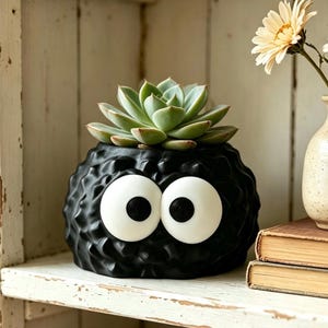 以下が含まれることがあります： 黒い質感のある丸いプランターで、黒い瞳を持つ2つの大きな白い円形の目が付いています。容器の中には緑色の多肉植物が植えられています。プランターの表面は凹凸のある不規則な仕上げになっており、毛羽立ったような外観をしています。