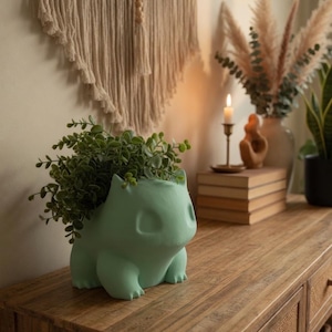 May include: A light green Bulbasaur-shaped planter filled with green foliage, sitting on a wooden surface. The planter is a stylized representation of the Pokémon character. The background includes a macrame wall hanging, a candle, and decorative plants.