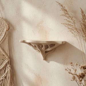Peut inclure: Une étagère murale dotée d'une finition beige clair lisse, avec une surface supérieure plate et un support décoratif en dessous qui imite la structure organique et ramifiée des racines d'arbres ou de branches entrelacées.