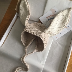 May include: A beige knitted baby hat with long bunny ears and a tie. The hat is in a white box with tissue paper.