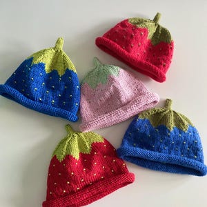 May include: A collection of handmade knit strawberry hats in various colors. The hats feature a red, pink, or blue body with green tops and small yellow bead accents, resembling strawberry seeds. The hats are designed for children.