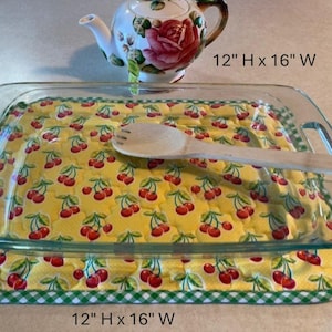 May include: A clear glass baking dish with a yellow cherry-patterned quilted cover, measuring 12" H x 16" W. A wooden spoon rests on the cover. A decorative teapot with a rose design is in the background.