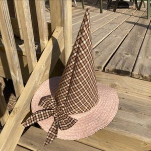 May include: A pointed hat featuring a tall, conical crown made of brown, black, and white plaid fabric. The wide, circular brim is covered in a textured, light pink material. A matching plaid fabric bow is attached to the base of the crown.