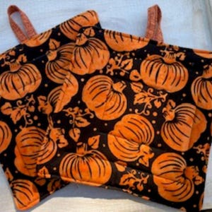 May include: Two square quilted pot holders featuring a repeating pattern of orange pumpkins and leaves on a black background. Each pot holder has a small orange fabric loop attached to the top corner for hanging, with visible straight-line stitching.