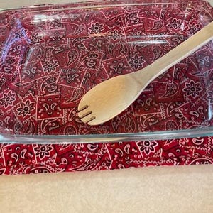 May include: A rectangular clear glass baking dish with integrated handles rests on a red and white fabric hot pad featuring a repeating paisley and floral bandana pattern. A light-colored wooden serving spoon with a slotted tip lies inside.
