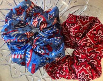 Puede incluir: Dos coleteros de tela. Uno presenta un fondo azul con un patrón patriótico de estrellas, rayas rojas y blancas y pequeños motivos de banderas estadounidenses. El segundo coletero tiene un estampado clásico de pañuelo de cachemira en rojo y blanco.