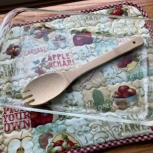 May include: A clear rectangular glass baking dish and a light-colored wooden serving spoon with a three-pronged fork end. The spoon rests inside the dish, which is placed on a quilted fabric mat featuring an apple orchard print with red apples.