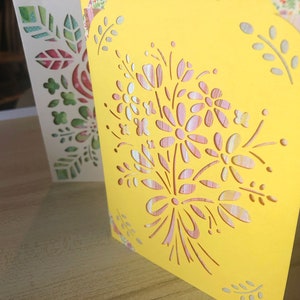 May include: A yellow card with a cut-out design of pink and white flowers and leaves. The card is decorated with a floral pattern.