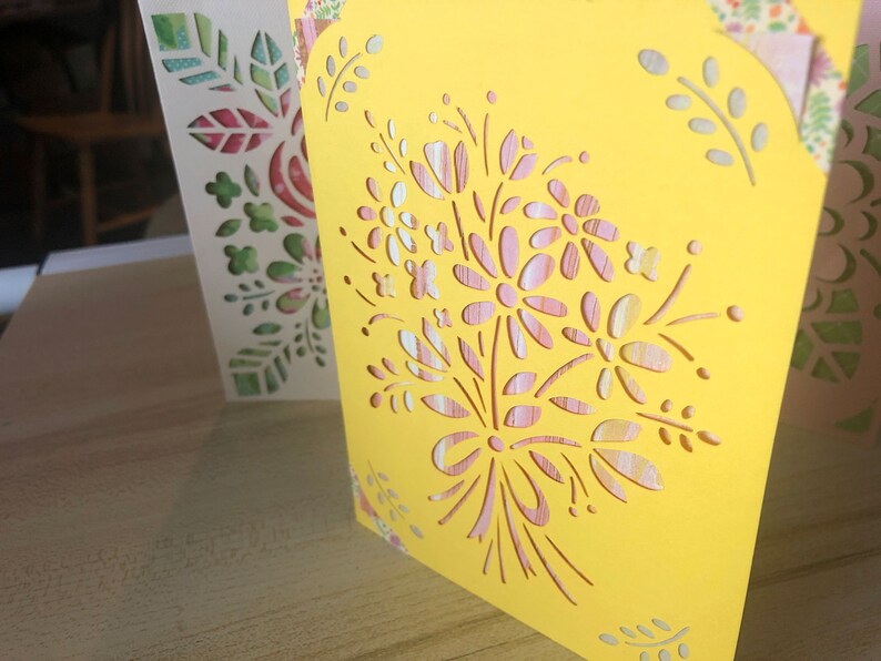 May include: A yellow card with a cut-out design of pink and white flowers and leaves. The card is decorated with a floral pattern.