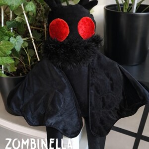 Puede incluir: Un peluche negro con forma de polilla con grandes ojos rojos y un cuello peludo. La polilla tiene grandes alas aterciopeladas y antenas. El texto "ZOMBINELLA ILLUSTRACIÓN PRINTS & APPAREL" está impreso en la parte inferior.