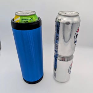 May include: A blue insulated tumbler with a black rim holding a green and yellow soda can. Two silver Pepsi cans are stacked next to the tumbler. The cans have the Pepsi logo and the words "Refresh, Recycle".