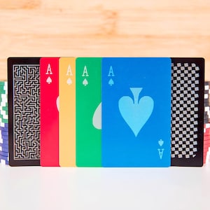 May include: Aces of spades playing cards in blue, green, yellow, and red, with a black card featuring a maze design. Stacks of poker chips in the background. The Ace of Spades card has a light blue spade symbol.