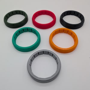 May include: A collection of six colorful, circular rings. The rings are in various colors, including green, red, black, teal, orange, and gray. Each ring has a textured interior. The rings are arranged on a white surface.