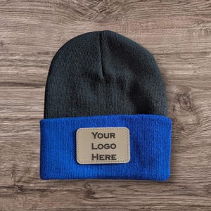 Puede incluir: Un gorro azul y gris oscuro con un parche rectangular que dice "YOUR LOGO HERE". El gorro está hecho de un material de punto y se muestra sobre una superficie de madera. La parte azul del gorro está doblada.