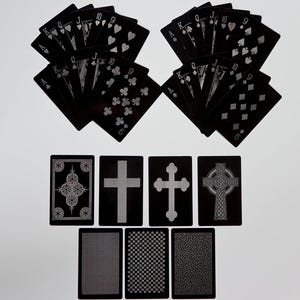 May include: A deck of black playing cards with silver suits and face cards. The image also shows several unique card backs with cross and geometric designs. The cards are arranged on a white surface.