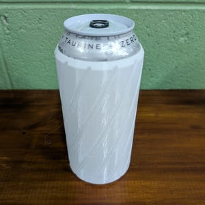 Pu&ograve; includere: Un portabevande bianco con un design a spirale, che contiene una lattina di bevanda. La lattina ha il testo "TAURINE ZERO" stampato sopra. La parola "White" &egrave; visualizzata nella parte inferiore dell'immagine.