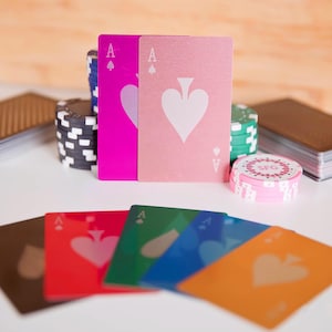 May include: A collection of poker playing cards and chips. The cards are in various colors, including pink, red, green, blue, and orange, each featuring a white spade symbol. Poker chips in stacks of black, green, and pink are also visible.