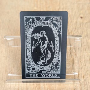 Puede incluir: Una carta de tarot negra que representa el Mundo. La carta tiene ilustraciones blancas de una figura sosteniendo dos varitas, rodeada por una corona. La carta se sostiene en un soporte de acrílico transparente. El texto "XXI" y "THE WORLD" son visibles.