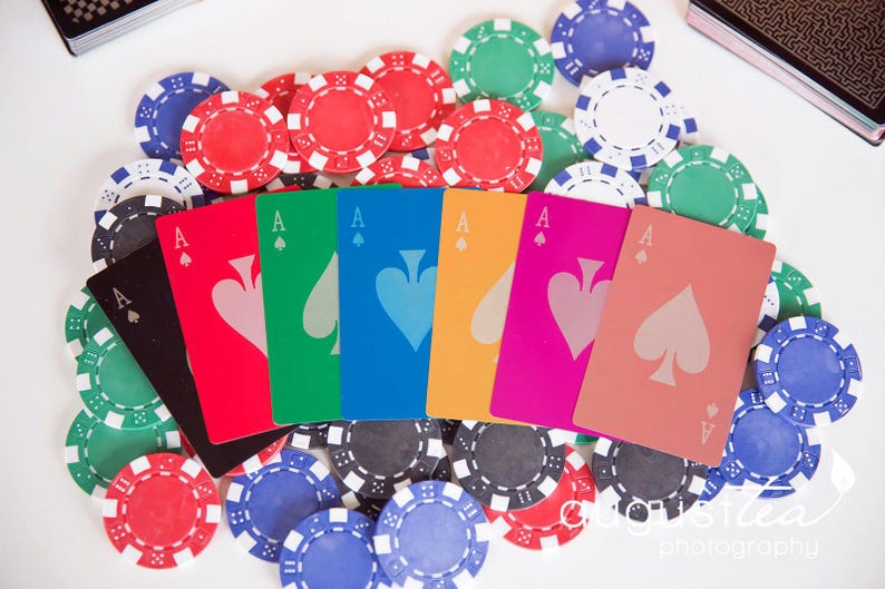 May include: A collection of poker chips in red, green, blue, and black surround a fan of playing cards. The cards are the Ace of Spades in various colors, including black, red, green, blue, yellow, pink, and peach.