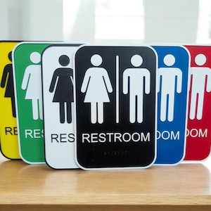 May include: A collection of restroom signs in various colors, including yellow, green, white, black, blue, and red. Each sign features a white stick figure icon and the word "RESTROOM" or "RES" in white text. The signs are arranged on a wooden surface.
