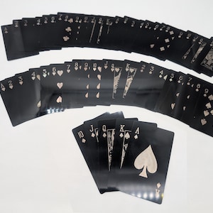 May include: A deck of playing cards with black card faces and light-colored suits and numbers. The cards are fanned out in multiple rows, with the suits of hearts, diamonds, clubs, and spades visible. The Ace of Spades is prominently displayed.