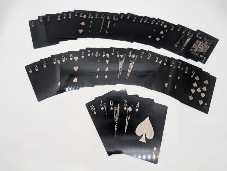 May include: A deck of playing cards with black card faces and light-colored suits and numbers. The cards are fanned out in multiple rows, with the suits of hearts, diamonds, clubs, and spades visible. The Ace of Spades is prominently displayed.