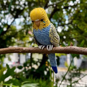May include: A crocheted budgerigar figurine featuring a bright yellow head, a light blue chest, and wings patterned with grey and yellow stitches. The bird has a tan beak, black eyes, and a long tail that transitions from light blue to dark blue.