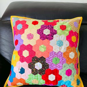 May include: A square pillow with a patchwork floral design. The pillow features a vibrant array of hexagon-shaped flowers in various colors, including red, pink, purple, and yellow. The pillow has a yellow border and is placed on a black surface.