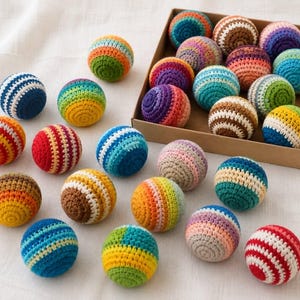 Op de afbeelding: Een verzameling kleurrijke, gestreepte haakballen in verschillende kleuren, waaronder blauw, rood, geel en groen. Sommige ballen zijn gerangschikt in een kartonnen doos, terwijl andere verspreid liggen op een wit oppervlak. Deze handgemaakte ballen zijn perfect om mee te spelen of te decoreren.