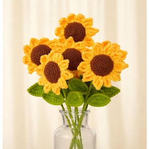 May include: A bouquet of crocheted sunflowers in a clear glass vase. The sunflowers have yellow petals and brown centers, with green leaves. The vase is filled with water, and the background is a light cream color.