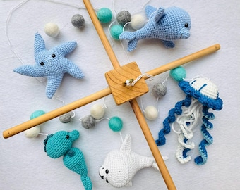 May include: A handmade baby mobile featuring crocheted sea creatures: a blue starfish, a dolphin, a seahorse, a seal, and a jellyfish. The mobile has a wooden frame and is decorated with felt balls in various colors.
