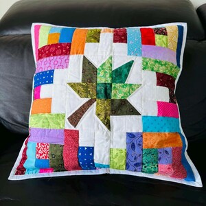 May include: Square patchwork pillow with a white star design at the center. The star is made of green, brown, and white fabric. The pillow's border is made of colorful fabric strips in various colors, including blue, orange, and pink.