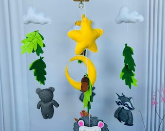 May include: A handmade baby mobile featuring felt clouds, leaves, a yellow star and crescent moon, a gray bear, a raccoon, and a hedgehog. The mobile is suspended from a wooden frame with a central hanging point.