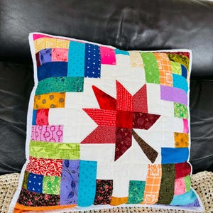 May include: Square patchwork pillow with a white background and a large red star in the center. The star is surrounded by a border of colorful fabric strips in various patterns and colors, including blue, green, and orange.