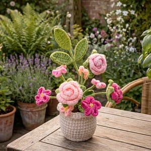 Puede incluir: Un arreglo floral de ganchillo en una maceta pequeña de color blanco roto. El arreglo presenta rosas rosas y otras flores rosas y fucsias con hojas verdes. La maceta está sobre una mesa de madera en un jardín.