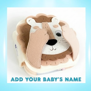 May include: A soft, fabric baby book shaped like a lion's face. The lion has a tan mane, light brown face, and tan ears. The text "ADD YOUR BABY'S NAME" is at the bottom of the image.