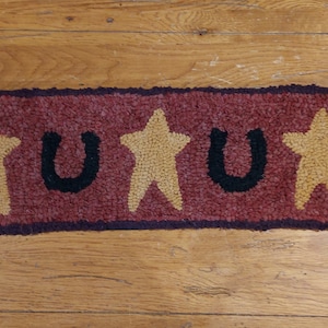 Puede incluir: Una alfombra de ganchillo roja, marrón y negra con dos herraduras negras y dos estrellas amarillas. La alfombra tiene un borde marrón oscuro.