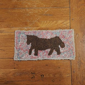 Op de afbeelding: Een klein, rechthoekig tapijt met een bruin silhouet van een paard op een roze, blauwe en witte achtergrond.