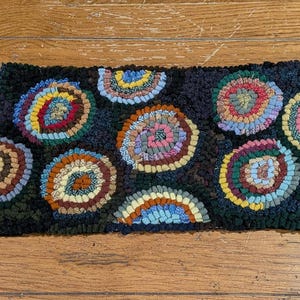 Puede incluir: Una alfombra rectangular cosida a mano, con un fondo oscuro y diseños circulares coloridos. Los círculos presentan varios colores, incluyendo rojo, amarillo, azul y verde. La alfombra está hecha de un material texturizado, probablemente lana o una tela similar.