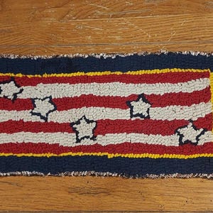 May include: A small, rectangular rug with a red, white, and blue American flag design. The rug is made of wool and has a fringed edge.