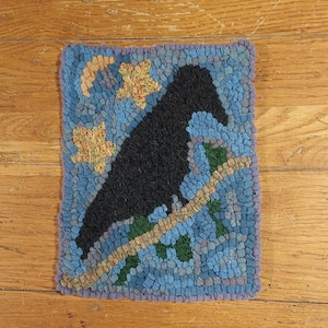 May include: A blue and green hooked rug with a black silhouette of a crow perched on a branch. The rug has a purple border and yellow flowers.