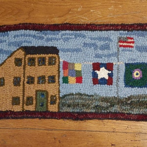 Puede incluir: Una alfombra hecha a mano que representa una casa marrón con ventanas y una puerta verde, un cielo azul con nubes y banderas, incluyendo la bandera estadounidense, la bandera de Texas, una bandera de colcha de retazos y una bandera verde con una flor morada.