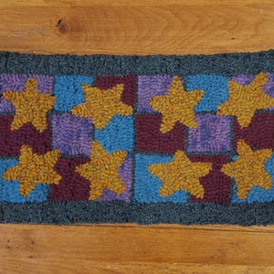 Puede incluir: Una alfombra hecha a mano con un diseño de patchwork que presenta un patrón repetitivo de cuadrados azules, morados y granates con estrellas amarillas en cada cuadrado.