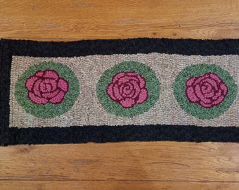 Puede incluir: Una alfombra rectangular con un borde negro y tres rosas rosas en círculos verdes sobre un fondo beige.