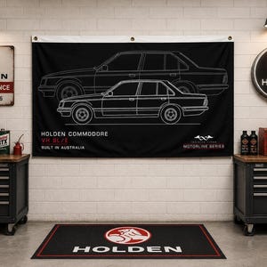May include: A black rectangular wall banner featuring a white line art illustration of a Holden Commodore VH SL/E car. The text reads HOLDEN COMMODORE VH SL/E BUILT IN AUSTRALIA and MOTORLINE SERIES. The banner has metal grommets at the top corners.
