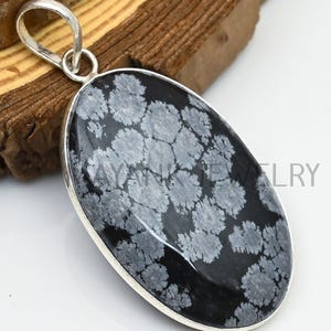Puede incluir: Colgante ovalado con una piedra de obsidiana copo de nieve negra y blanca, engastada en un marco de metal plateado. El colgante tiene un lazo para una cadena. El texto "MAYANK JEWELRY" es visible en la parte superior de la imagen.