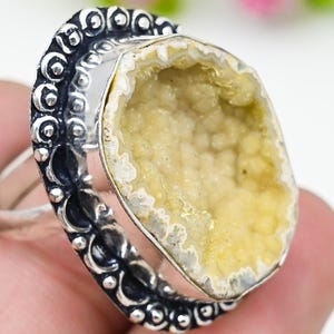 May include: A silver ring featuring a large, oval-shaped, light yellow geode stone. The stone is set in a silver bezel and surrounded by a decorative, dark silver band with intricate detailing. The ring is displayed against a soft, blurred background.
