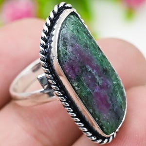 Op de afbeelding: Een zilveren ring met een grote, rechthoekige edelsteen met een groen en kastanjebruin patroon. De edelsteen is gezet in een zilveren bezel met een gedraaid touwontwerp. De ring zit aan een vinger.