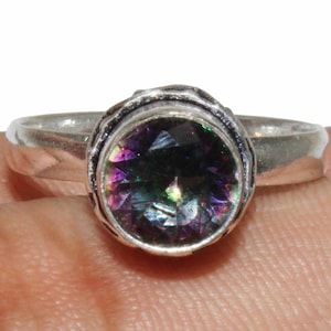 Może przedstawiać: Srebrny pierścionek z okrągłym, tęczowym kamieniem szlachetnym osadzonym w srebrnej oprawie. Kamień szlachetny prezentuje tęczę kolorów, w tym fioletowy, zielony i niebieski.