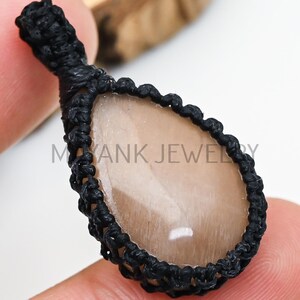 May include: A teardrop-shaped pendant with a light brown stone, set in a black macrame weave. The pendant is held by a finger. The text "MAYANK JEWELRY" is visible at the top and bottom of the image.